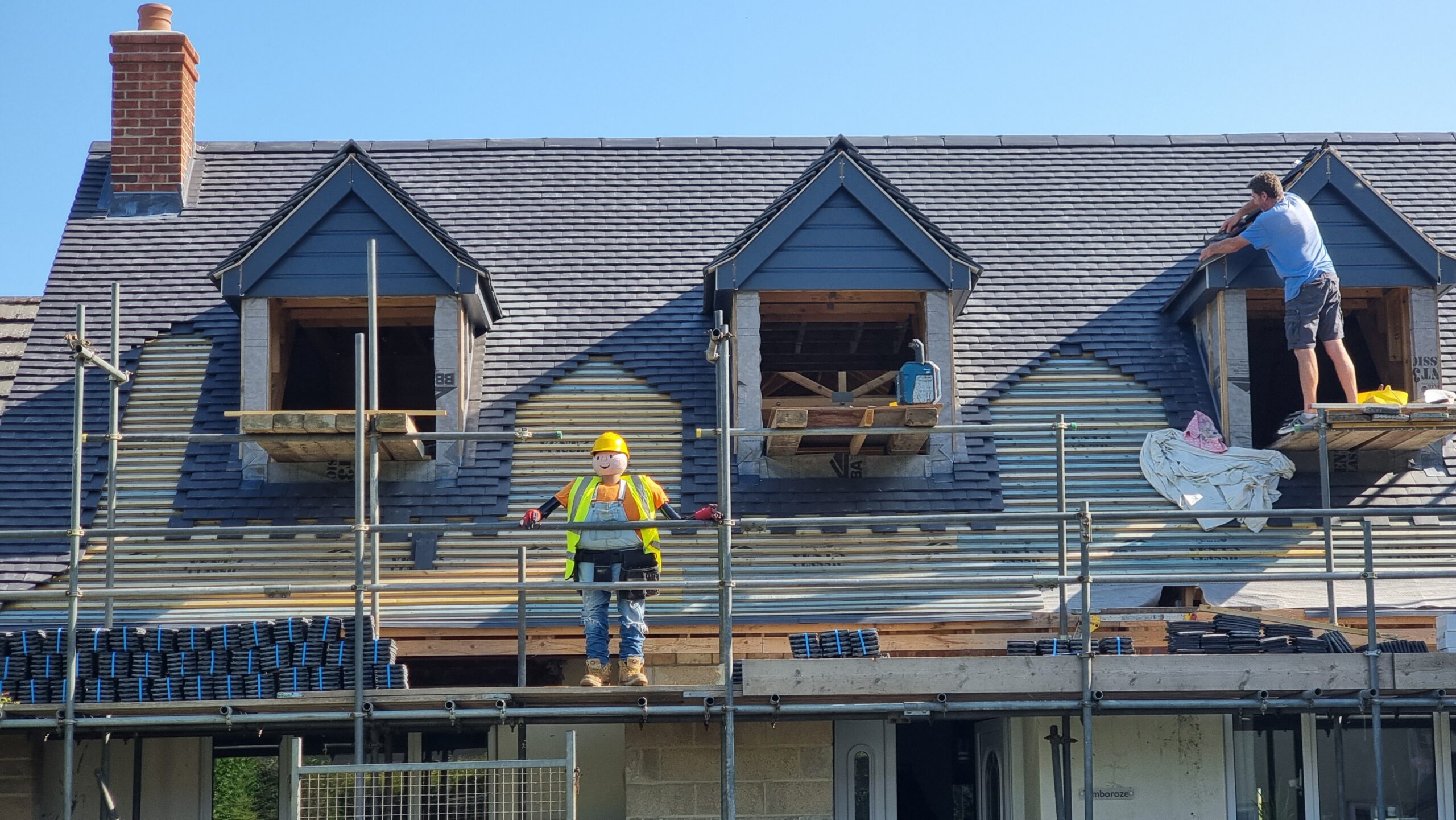 Full image of A lifesize mannequin of kids' TV character Bob the Builder stands on scaffolding that's being used by an actual builder who's constructing hip-roof dormer windows.