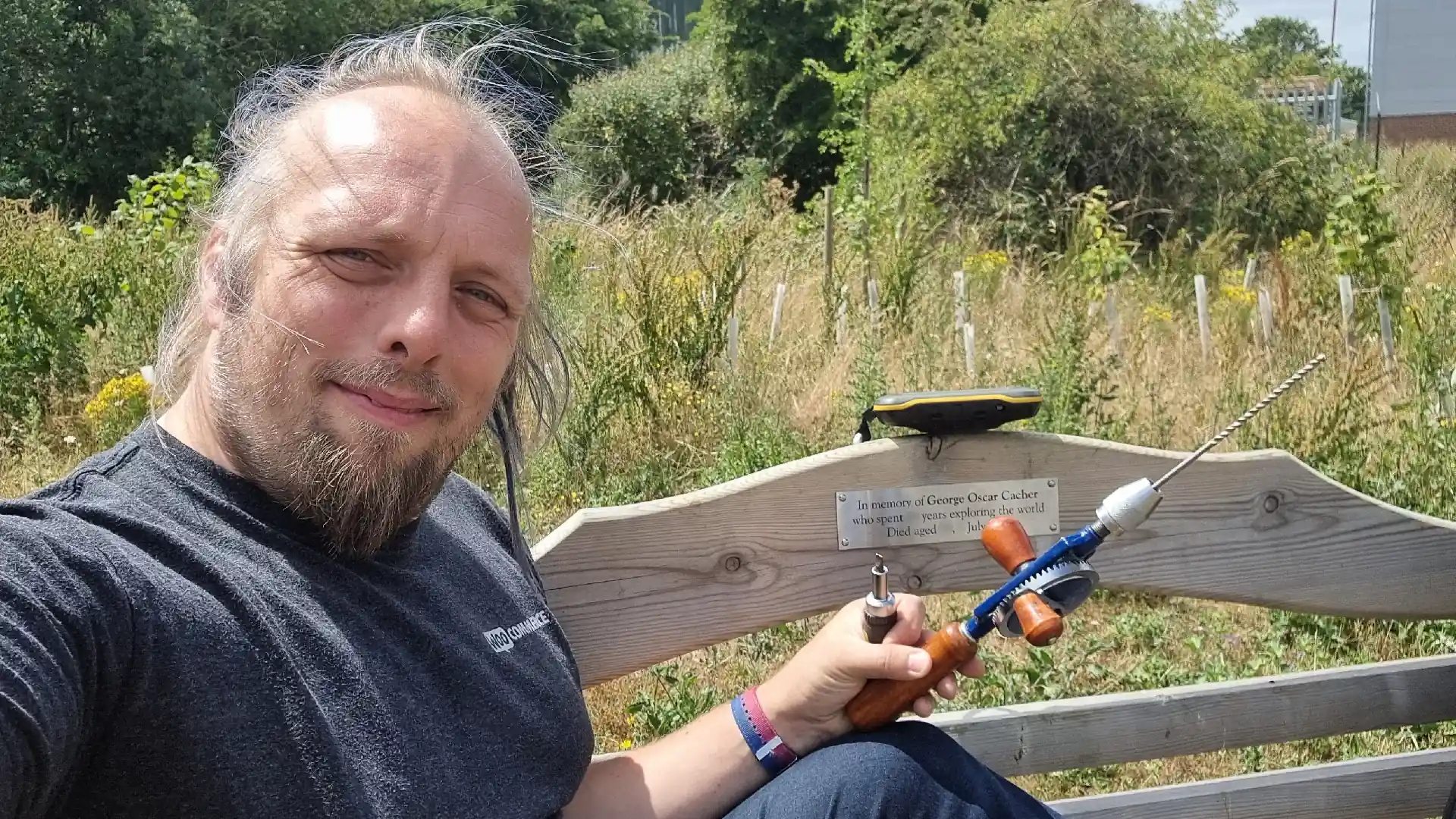 Full image of Dan, a white man with a beard and long, blue-tinted hair, sits sideways on a wooden bench behind which overgrown tree planting can be seen. On the back of the bench is perched a handheld GPS receiver, and in Dan's hand is a small screwdriver and a hand drill. The bench has a plaque attached (parts of which have been censored for the photo), which reads: In memory of George Oscar Cacher who spent ... years exploring the world. Died aged ..., ... July ....