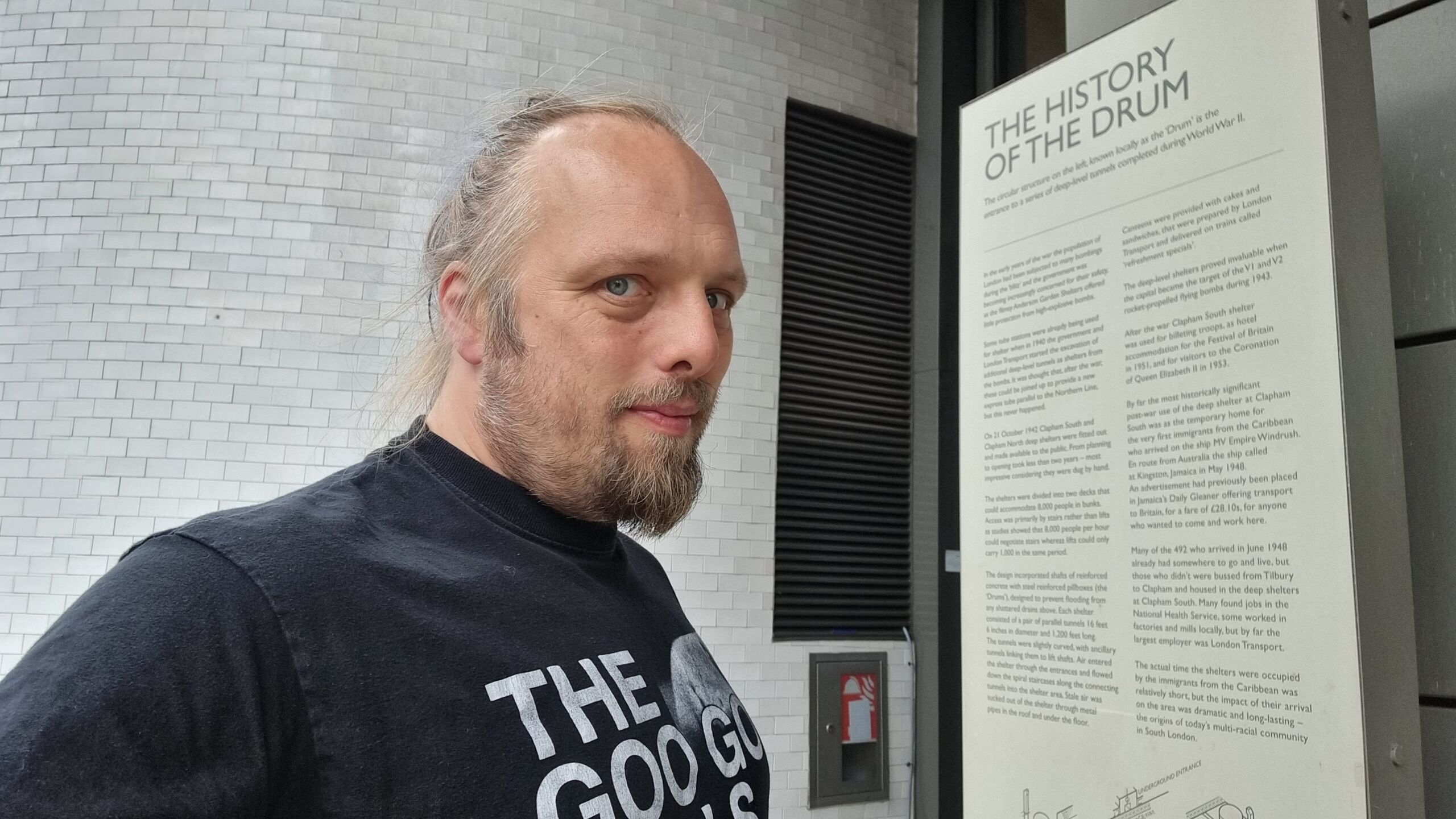 Full image of Dan, a white man wearing a Goo Goo Dolls t-shirt, stands in front of a sign describing the history of The Drum in South Clapham.