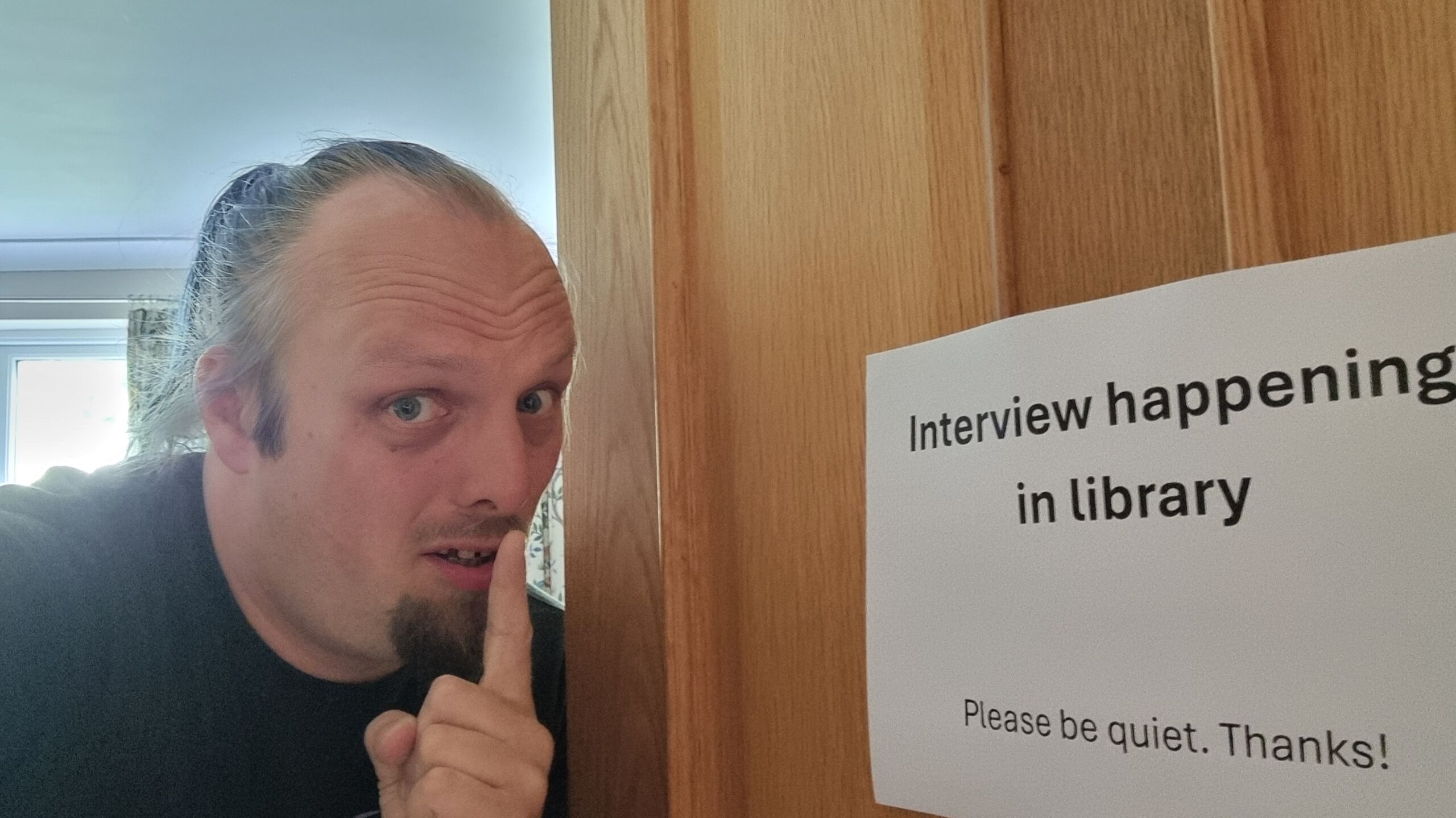 Full image of Dan peeps out from behind a door, making a 'shush' sign with his finger near his lips. On the door is a printed sign that reads 'Interview happening in library. Please be quiet. Thanks!'
