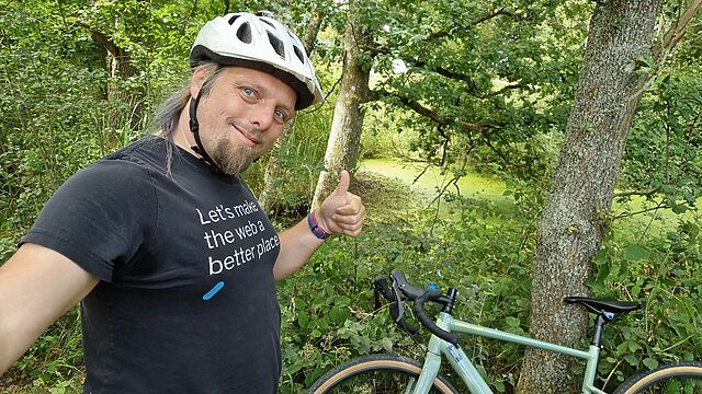 Dan, wearing a cycle helmet, throws a thumbs-up to the camera from a densely-wooded path.