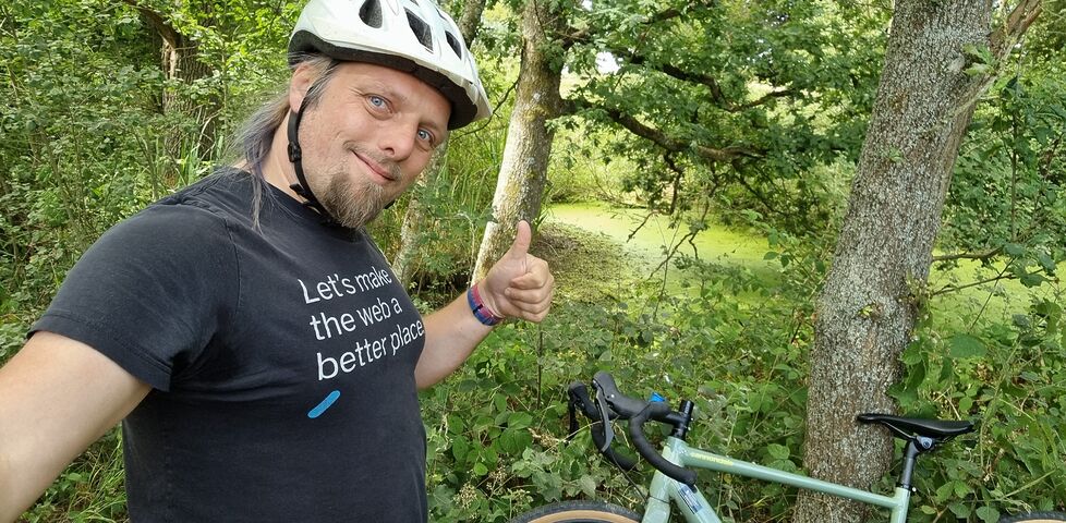 Dan, wearing a cycle helmet, throws a thumbs-up to the camera from a densely-wooded path.