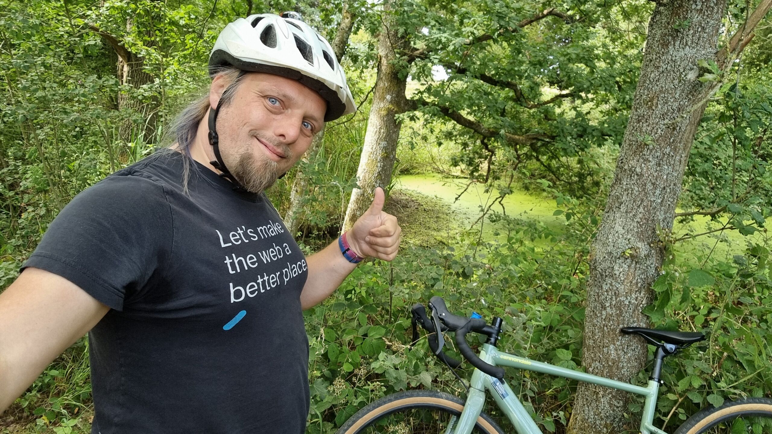 Full image of Dan, wearing a cycle helmet, throws a thumbs-up to the camera from a densely-wooded path.