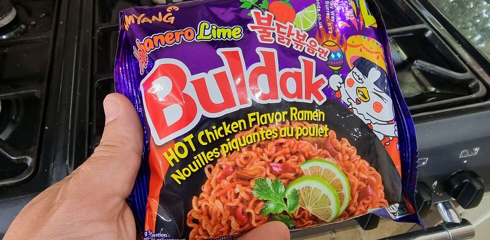 In a kitchen, a hand holds a purple foil packet of Samyang Buldak noodles, "Habenero Lime" flavour.