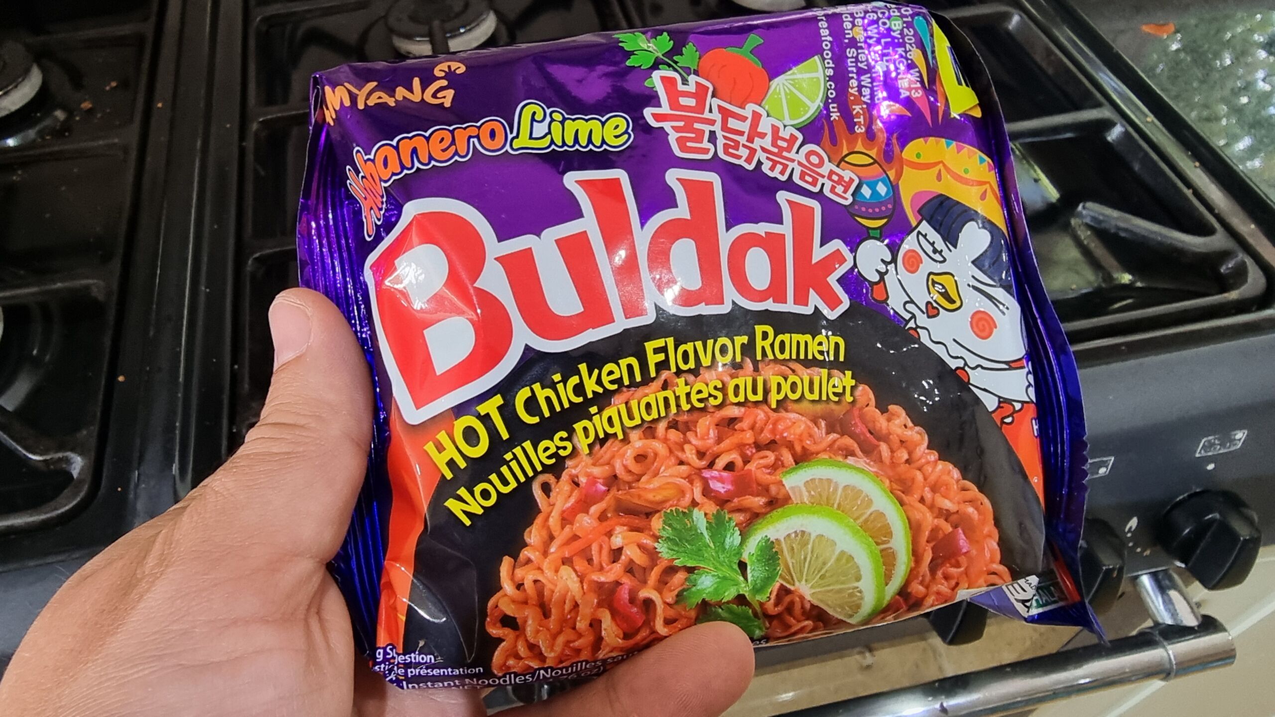 Full image of In a kitchen, a hand holds a purple foil packet of Samyang Buldak noodles, "Habenero Lime" flavour.