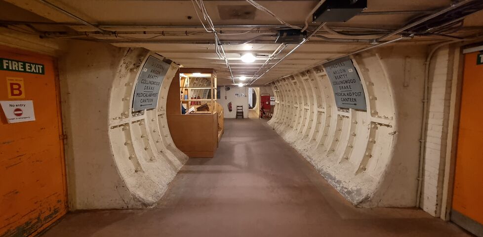 A circular tunnel with a flat floor and low ceiling, wrapped in white-painted iron and concrete, with exposed wiring and WWII-era signage, leads deeper into a subterranean bunker past a small guard post in which a figure can barely be seen, reading a book.