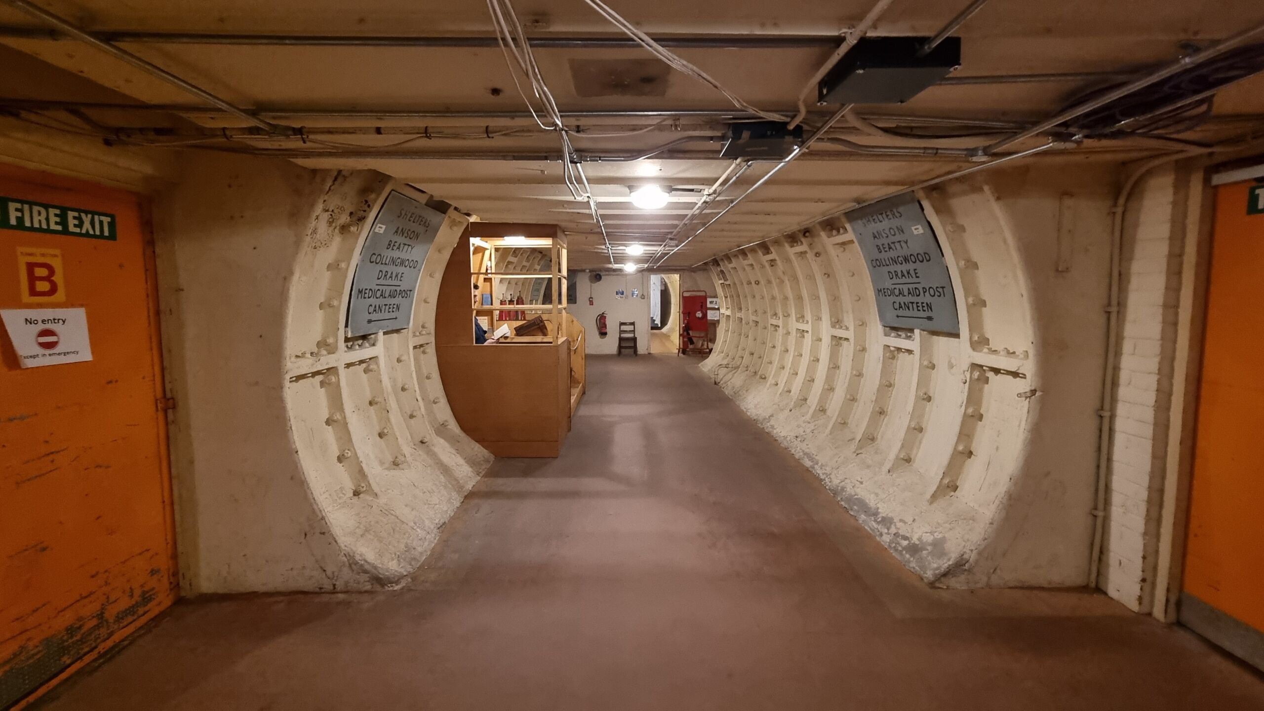 Full image of A circular tunnel with a flat floor and low ceiling, wrapped in white-painted iron and concrete, with exposed wiring and WWII-era signage, leads deeper into a subterranean bunker past a small guard post in which a figure can barely be seen, reading a book.