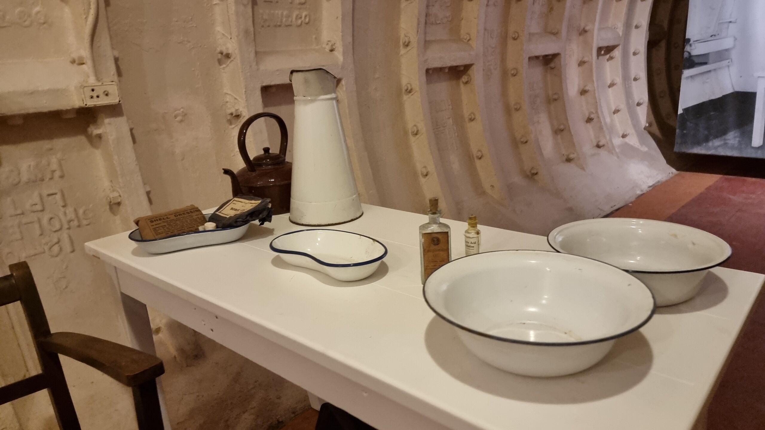 Full image of Table with basins, kidney bowl, old-style medical vials, kettle, jug, and WWII-era shrapnel dressing kit, in an iron-framed subterranean tunnel.