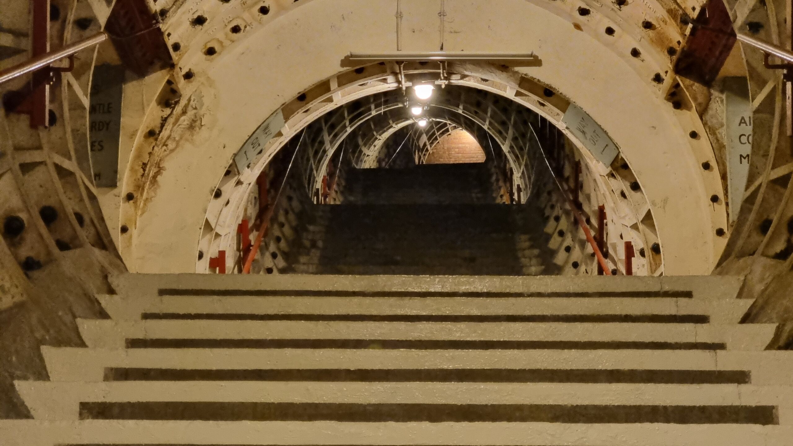 Full image of Broad staircase reminiscent of those found around the London Underground, but sparsely lit and decorated. Looking up the stairs, it ends in a brick wall.