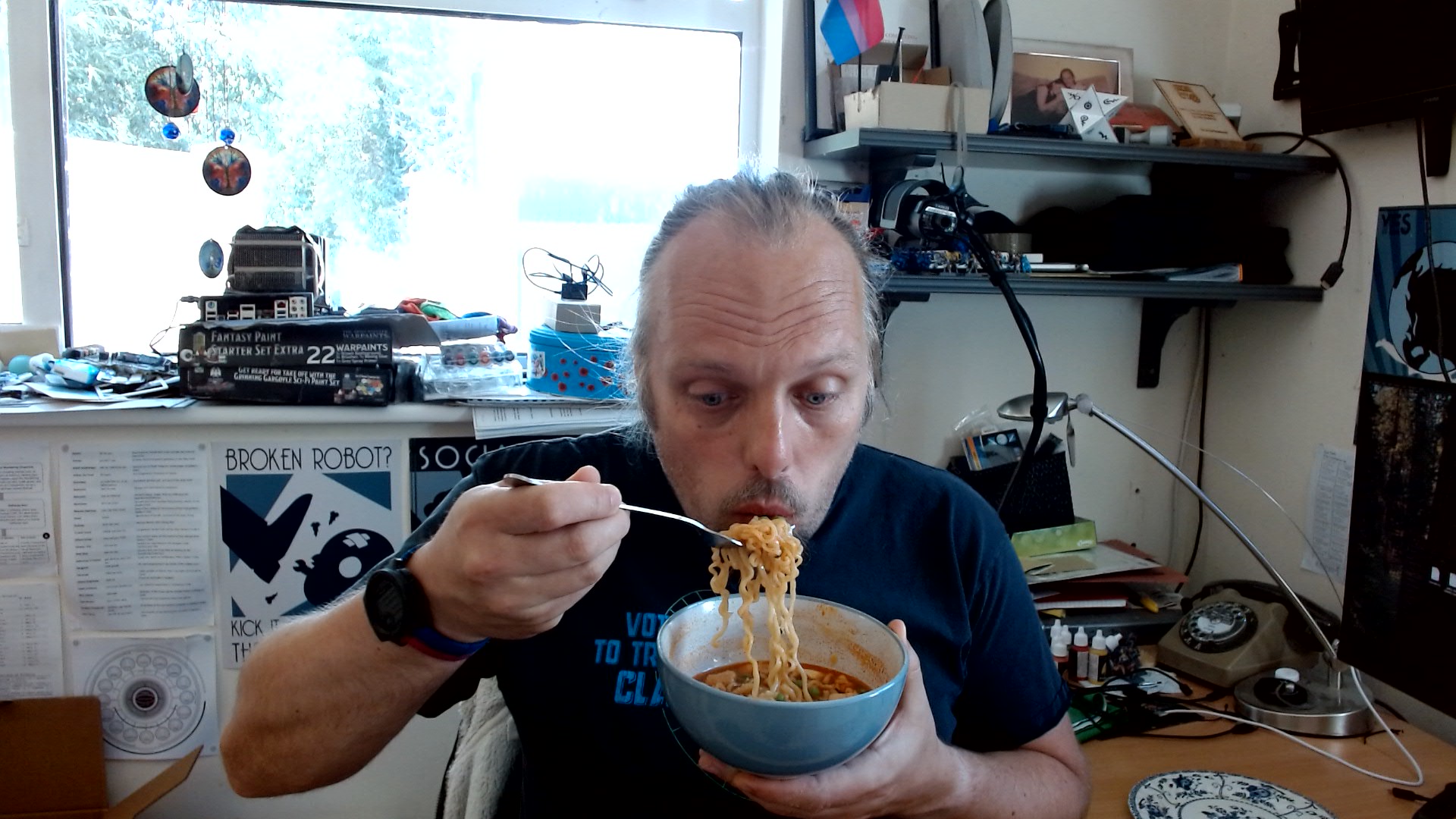 Full image of Dan slurps a forkful of noodles lifted from a bowl full of noodles in a deep red sauce, in a cluttered office space.