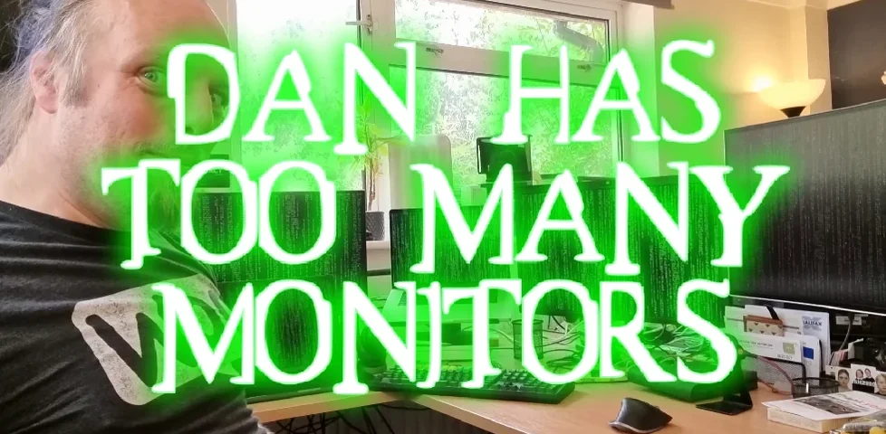 Photo of Dan, smiling, in front of a row of monitors showing falling green code in the style of The Matrix. Overlaid, in a Matrix font, green text reads 'Dan Has Too Many Monitors'.