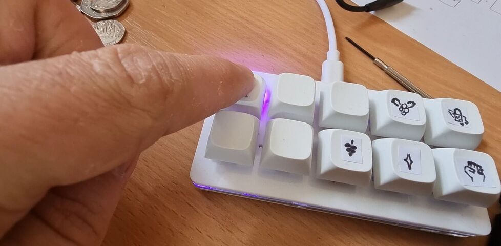 A small white 10-key keyboard with hand-drawn icons on stickers on the keys. A finger reaches in to press one key, which flashes a purple LED from beneath.