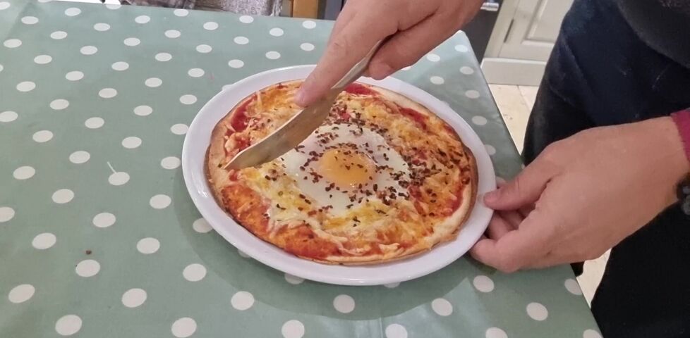 On a white plate on a kitchen table, a knife is about to make a first cut to slice a quesapizza that's topped with tomato, cheese, a baked egg, and chilli flakes.