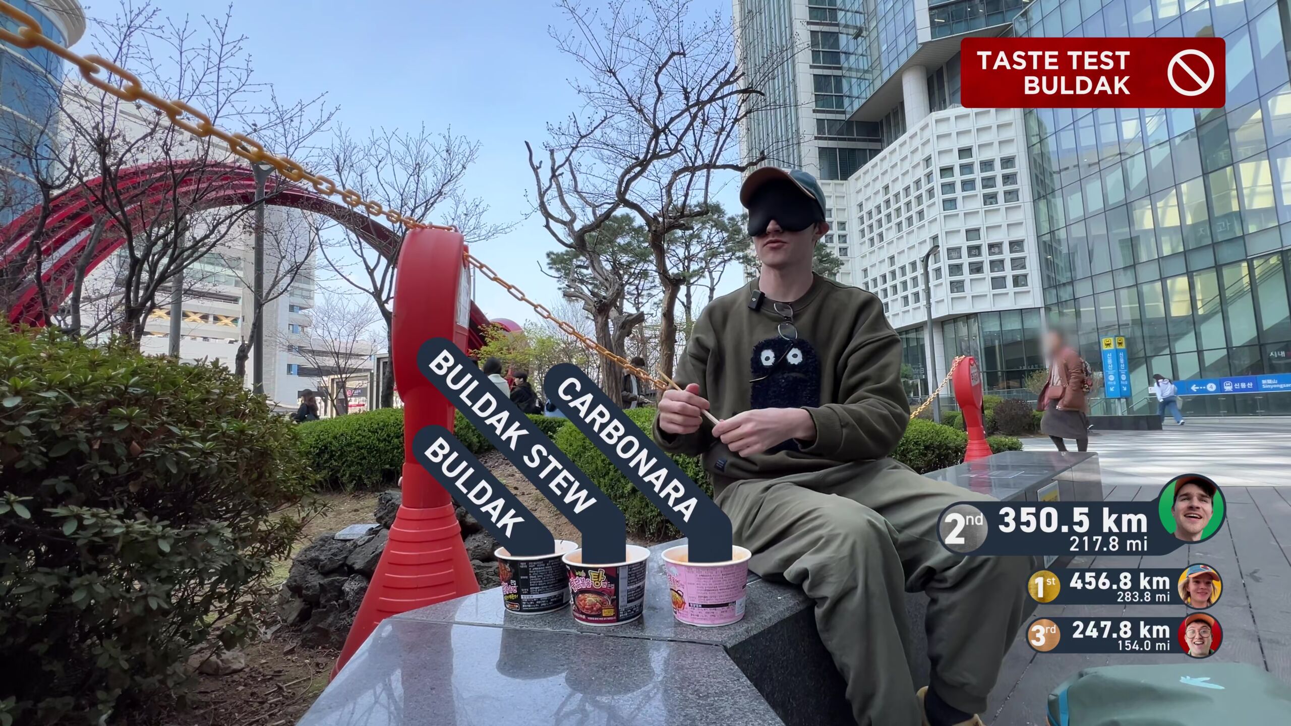 Full image of Framegrab from Jet Lag: The Game, showing Adam Chase, a young white man, sitting in a South Korean urban centre, blindfolded, holding chopsticks and preparing to eat from three bowls of instant noodles, captioned Buldak, Buldak Stew, and Carbonara.
