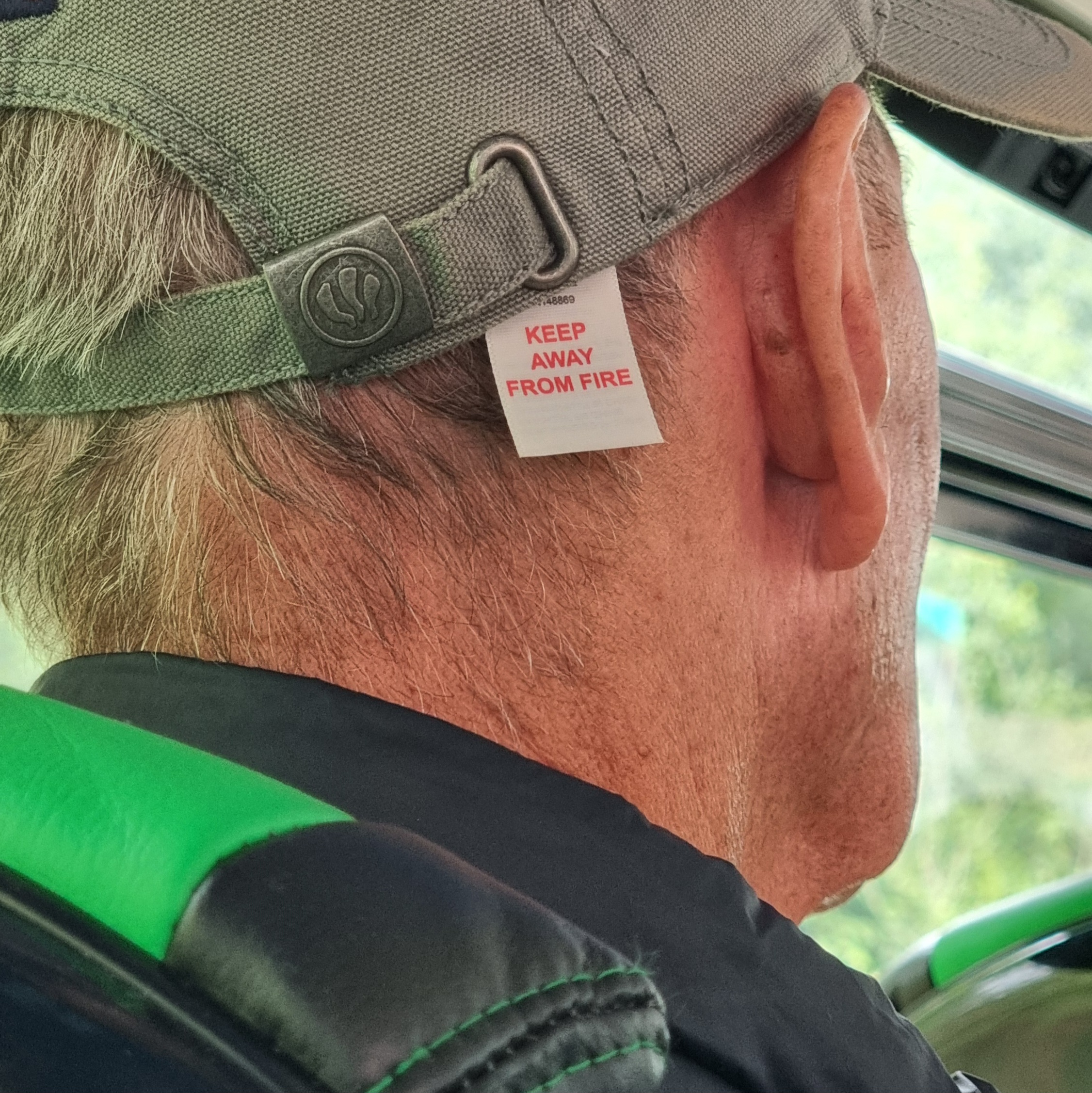 Full image of On a bus, back abs sides of a man's head. His hat has a label which reads "keep away from fire", but it's hanging out in such a way that it looks like it might be his head that's labelled.