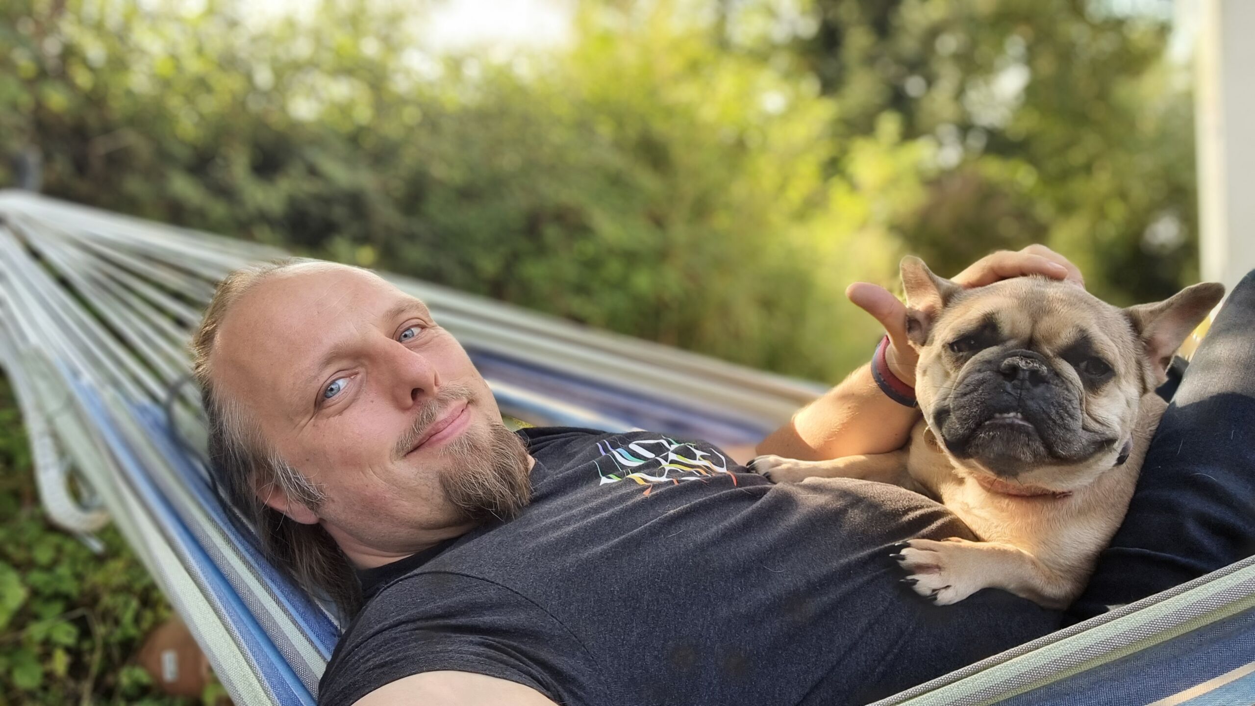 Full image of Dan, a middle-aged white man with long hair and a goatee beard, lies on a striped hammock in a verdant garden. He's wearing a t-shirt which says "love" over a ribbon in pride colours, and on his chest lies a contented-looking champagne-coloured French Bulldog.