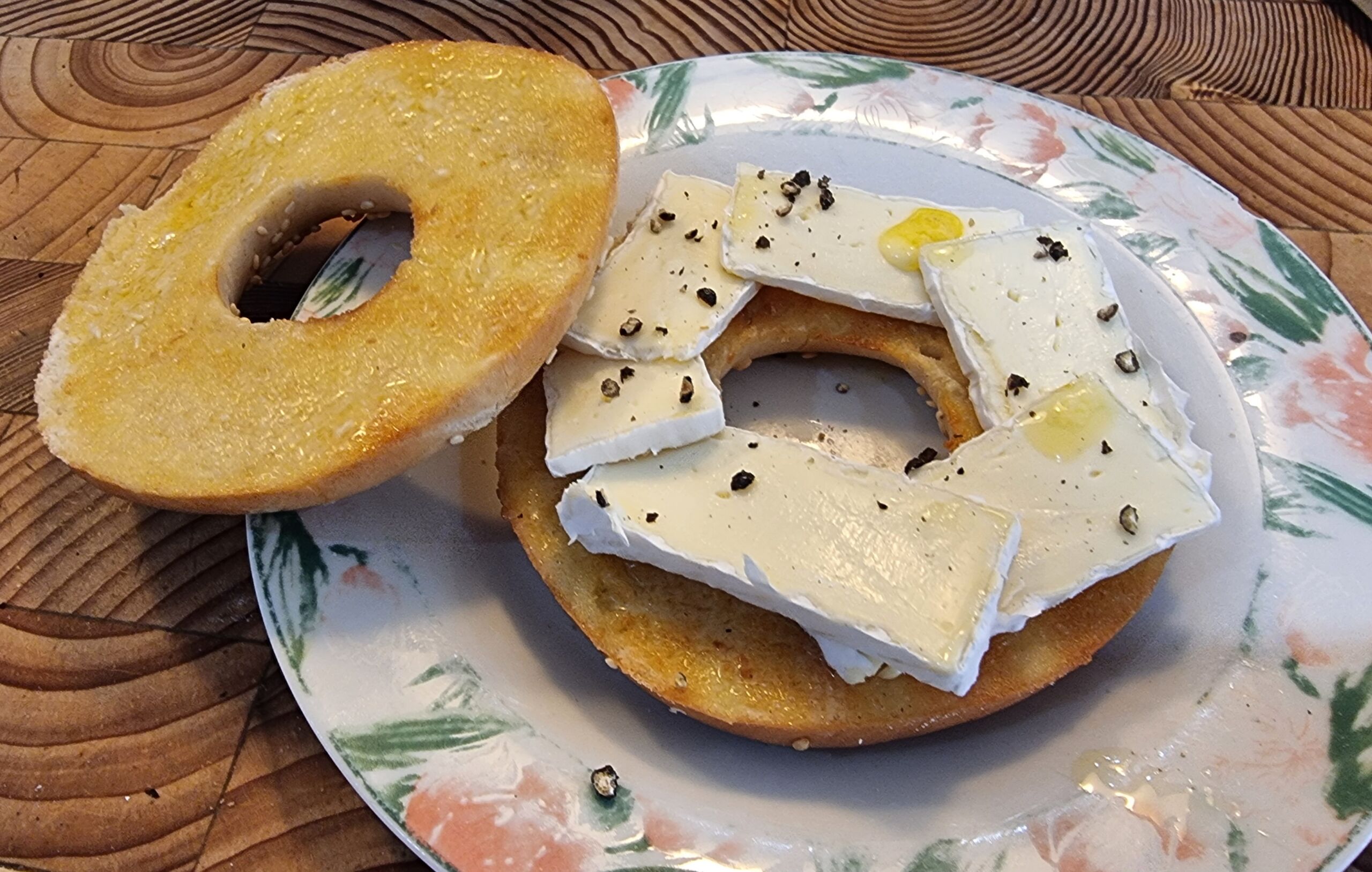 Full image of On a floral plate atop a wooden surface z an open sesame bagel contains an Escherian 'endless staircase' of sliced brie.
