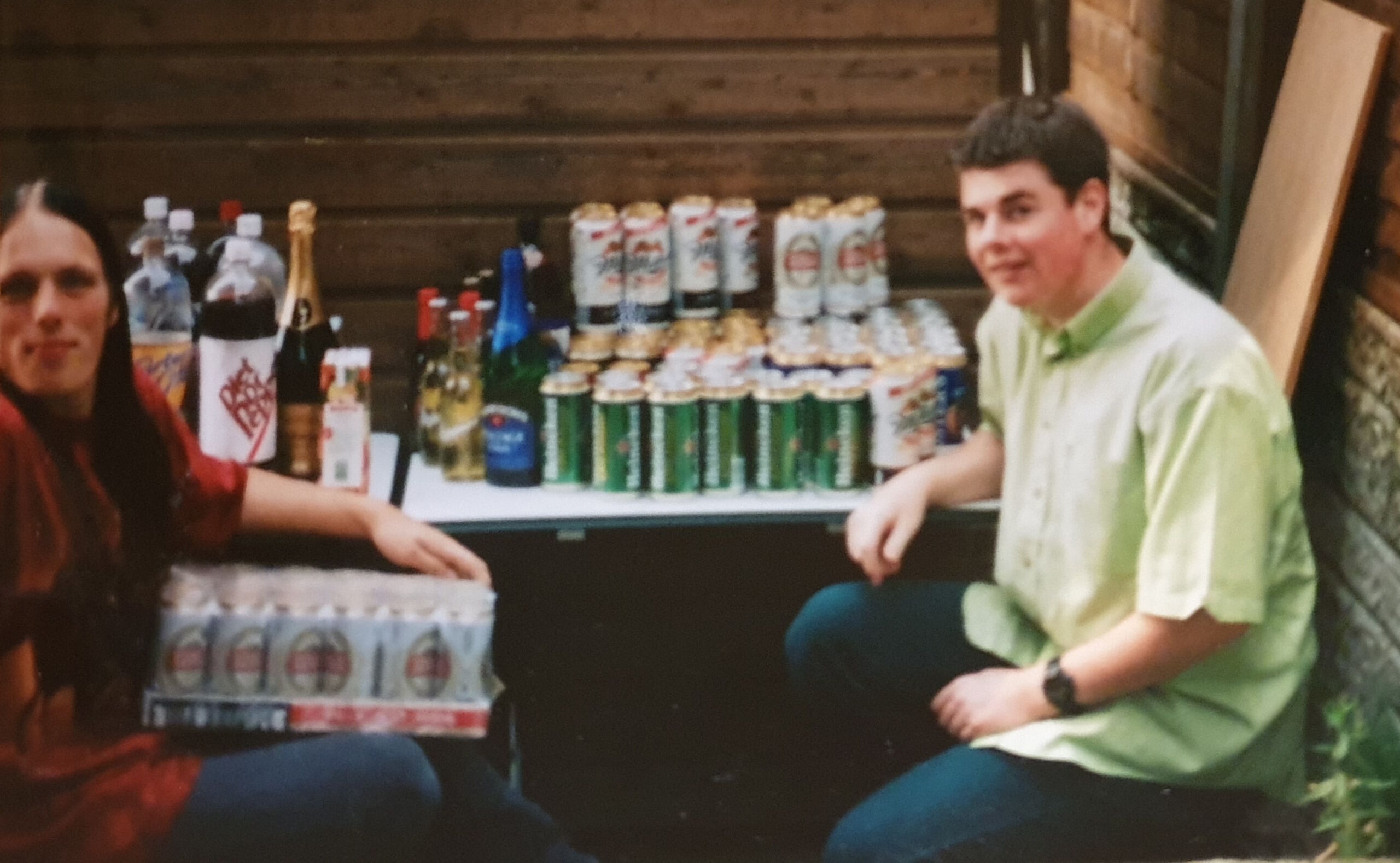 Full image of Dan, then with long black hair, sits with another young white man in a garden, in front of a table laden with several bottles of wine and around 80 cans of beer.