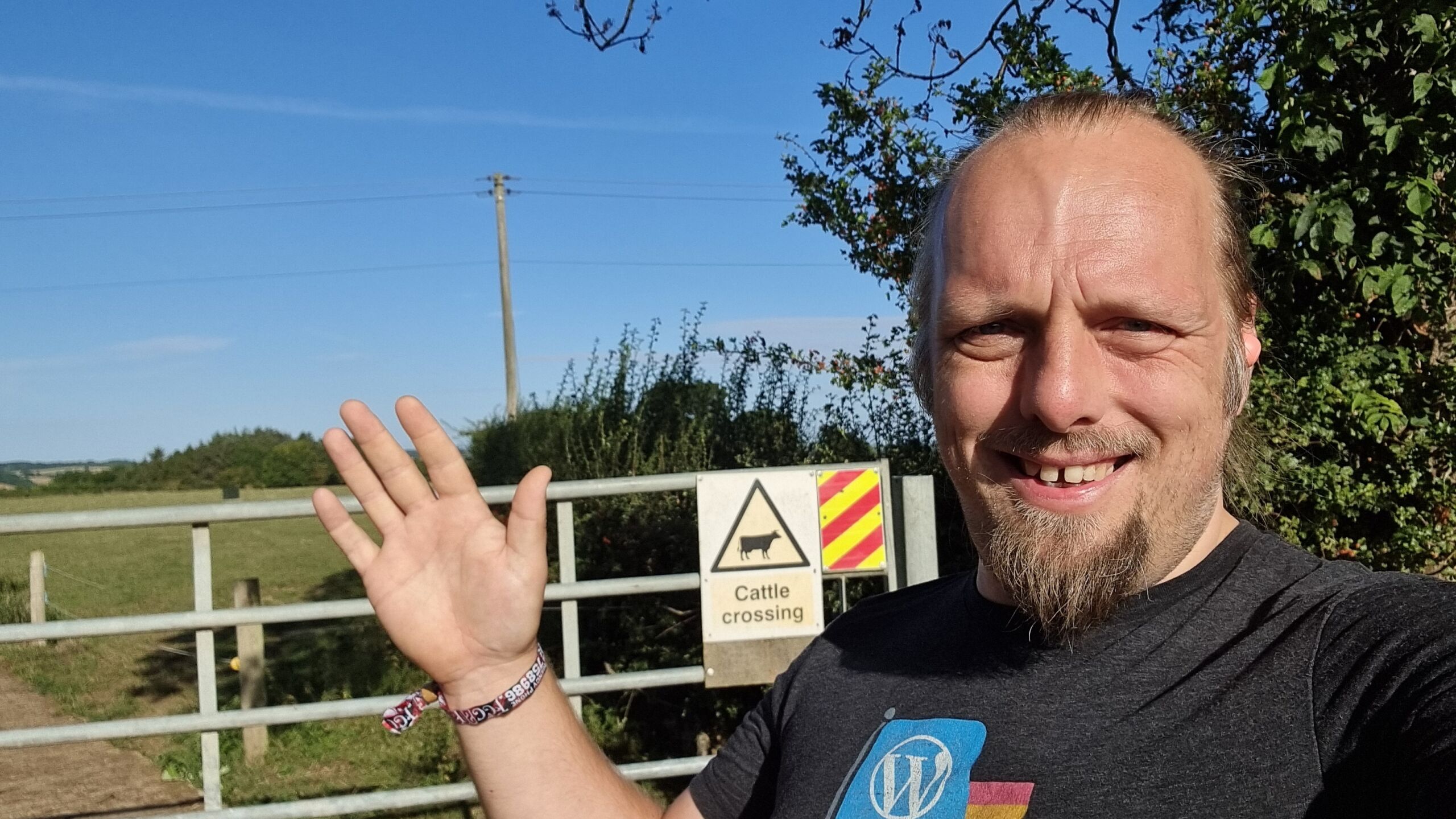 Full image of In front of a gate with a 'cattle crossing' sign, Dan waves to the camera with a hand whose wrist has a Cropredy 2025 wristband.