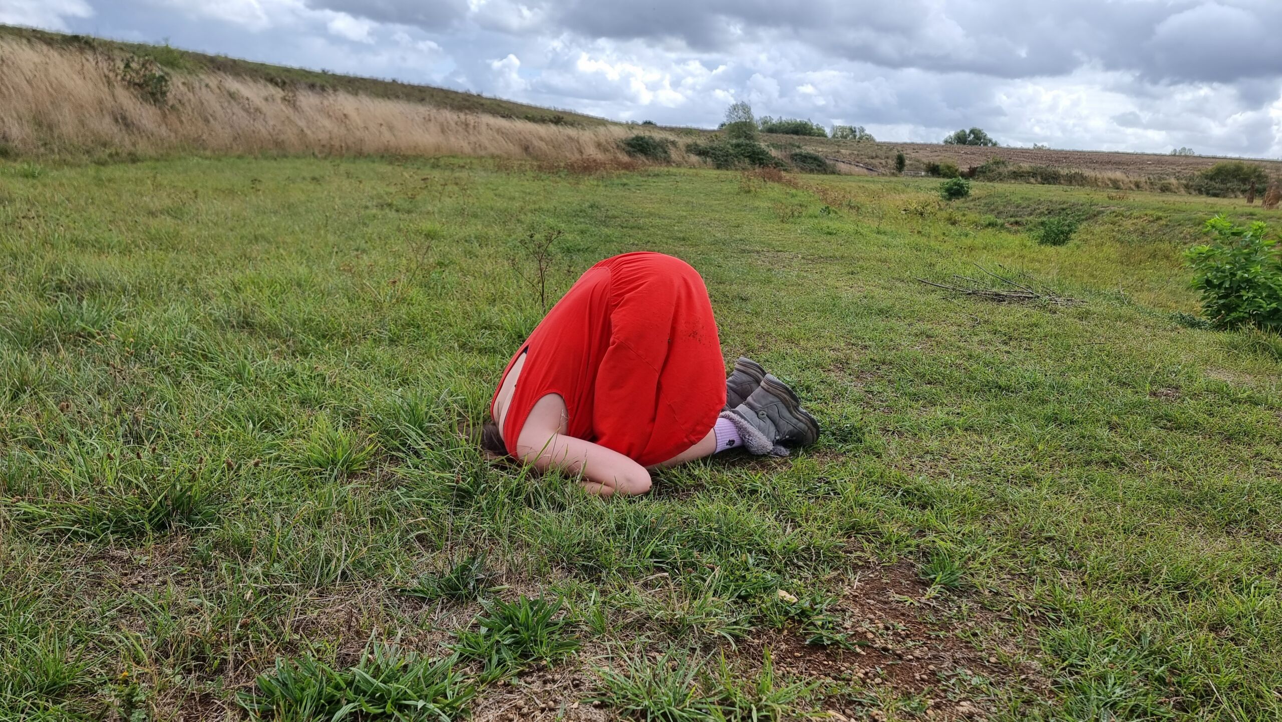 Full image of In a grassy field, a girl in a red dress and comfortable boots kneels with her head completely vanished down a rabbit hole.