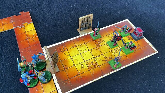 Several jigsaw-edged board game pieces lay out a dungeon map, with painted plastic minatures representing doors and characters. A party of four adventurers have just opened a door into a chamber containing five skaven (ratmen), guarding a treasure chest.