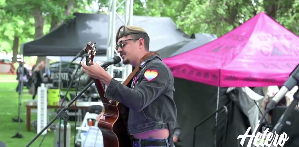 A man wearing an army beret and a jacket with a pride-rainbow heart on the sleeve plays acoustic guitar on a festival stage.