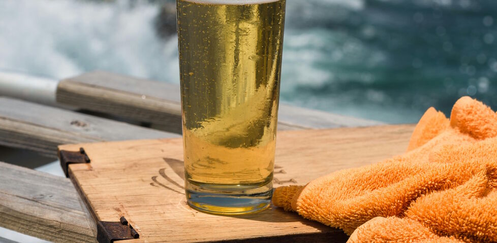 A pint of light-coloured beer on wooden board, with the sea in the background. An orange towel sits alongside.