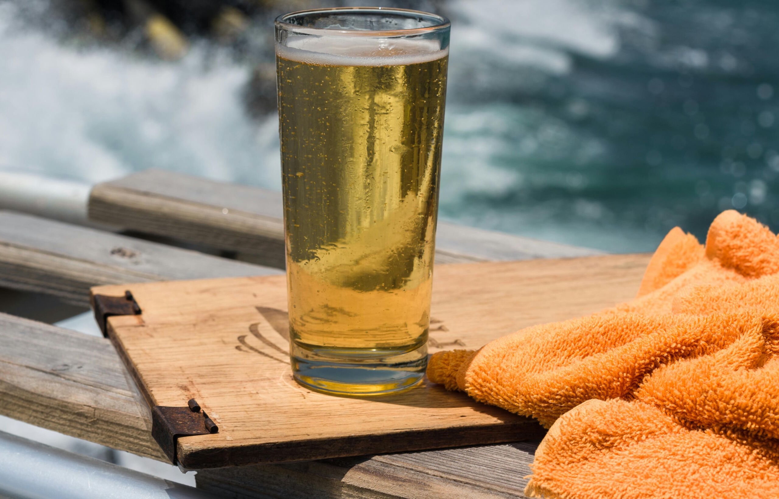 Full image of A pint of light-coloured beer on wooden board, with the sea in the background. An orange towel sits alongside.