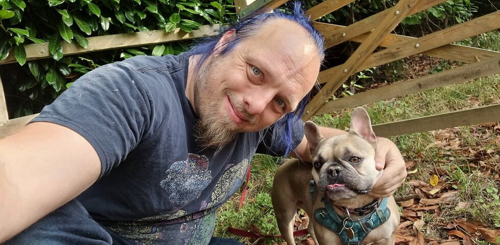 Dan, a white man with blue hair tied in a ponytail and a goatee beard, crouches alongside and pets a champagne-coloured French Bulldog by a gate on a path through a managed forest. There are bits of plant on his clothes, especially his shoulders.