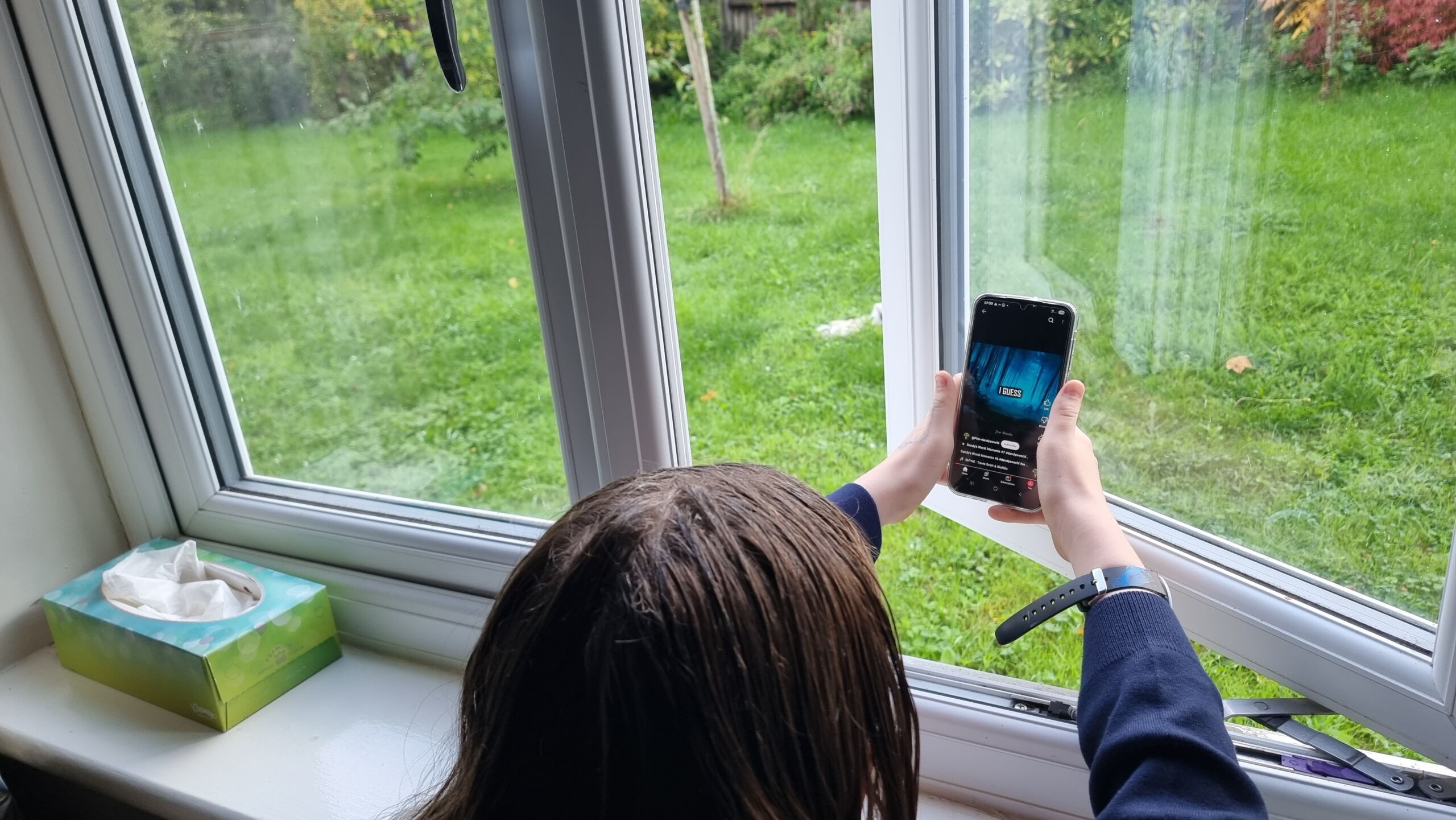 Full image of A child leans out of an open window into a rainy garden, holding her phone outdoors to watch a YouTube video which shows the words 'I GUESS' over a blue-tinted forest background.