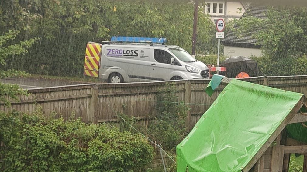 Full image of A silver van branded 'Zero Loss' has its doors open in a rainy road, as seen from the window of a nearby house, over its garden.