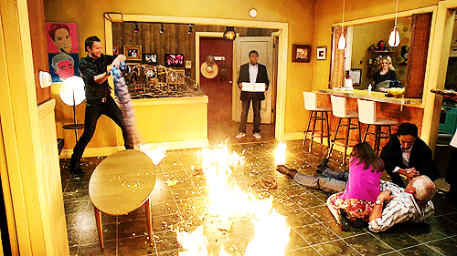 Scene from Community episode 'Remedial Chaos Theory'. Pierce lies injured on the floor, tended to by Annie and Abed, while Jeff swings a flaming blanket around his head. Troy stands in shock at the door, holding a pile of pizza boxes.