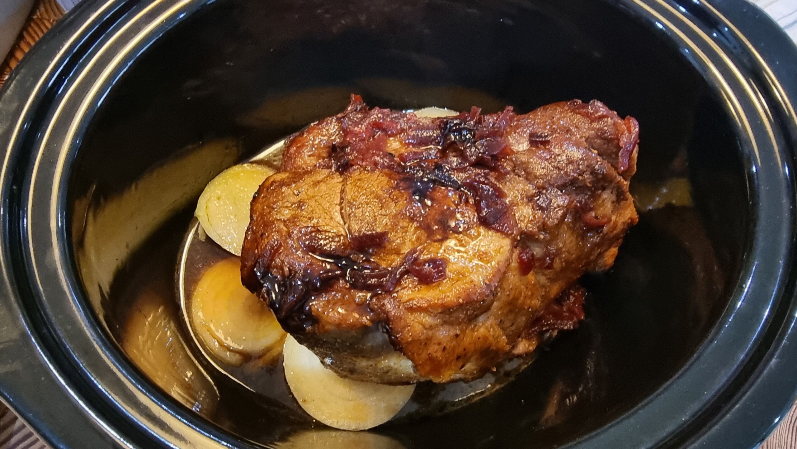 Full image of A seared shoulder of pork rubbed with spices sits in a syrupy cider mix atop some sliced white onions in a slow cooker pot.