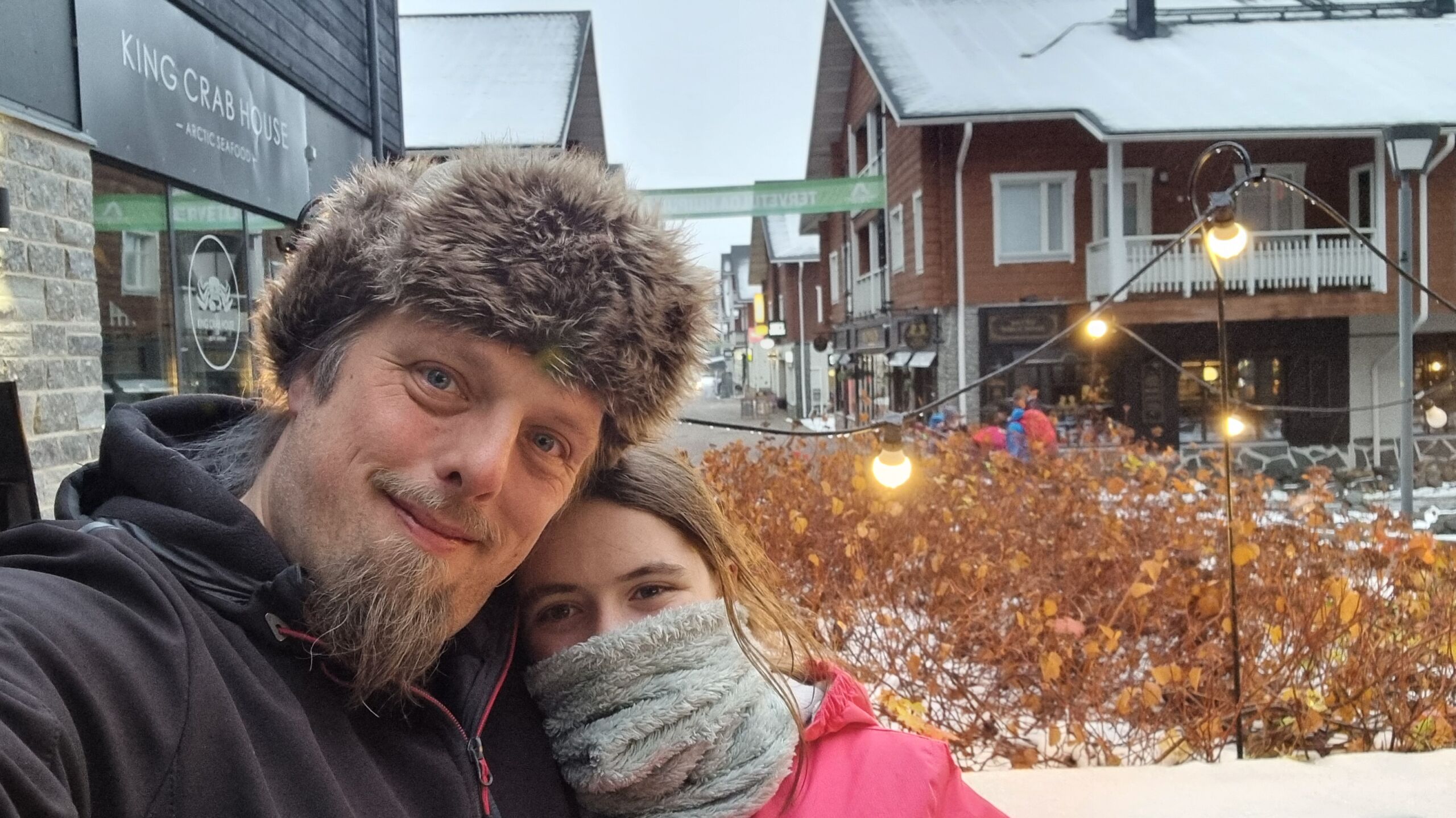 Full image of Wrapped up in warm hats and scarves, Dan and a tweenage girl stand in the snowy street of a Lapland town.