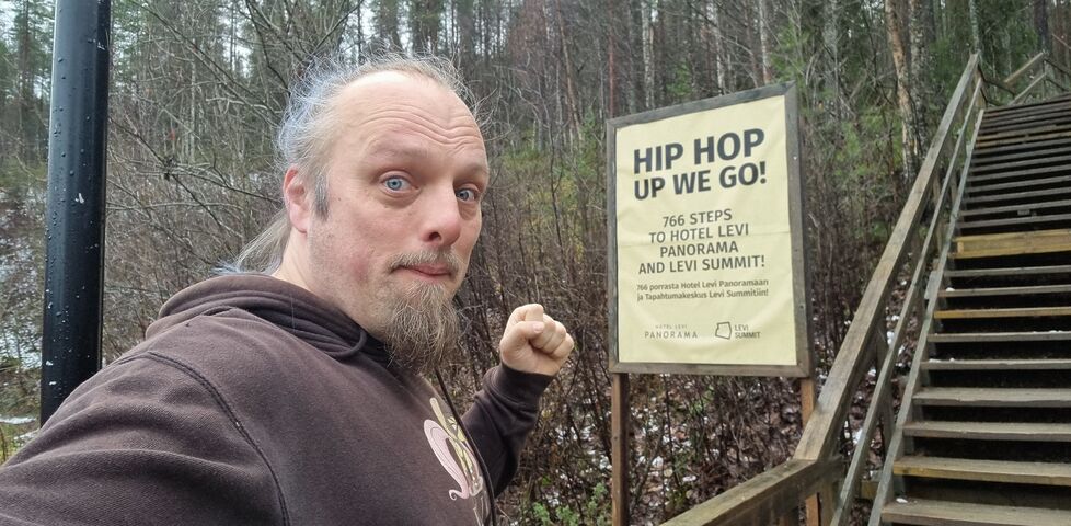 Dan prepares to jog up a wooden hillside staircase, marked with a sign reading: 'Hip Hop, Up We Go! 766 steps to Hotel Levi Panorama and Levi Summit'