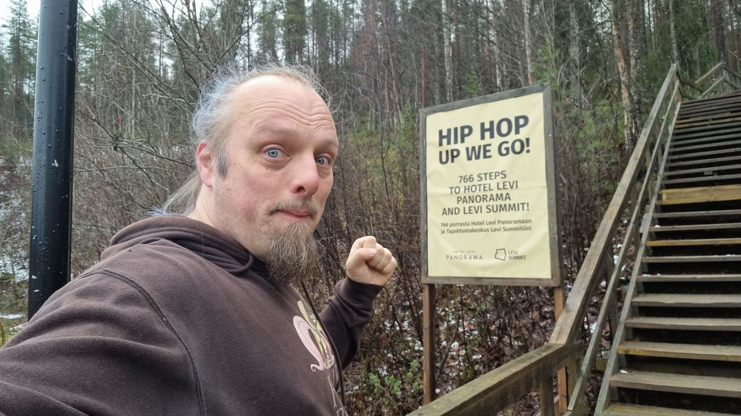 Full image of Dan prepares to jog up a wooden hillside staircase, marked with a sign reading: 'Hip Hop, Up We Go! 766 steps to Hotel Levi Panorama and Levi Summit'