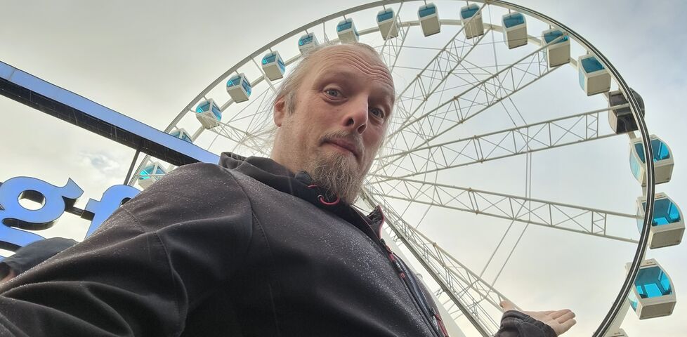 Dan stands in front of a ferris wheel against a cloudy sky.