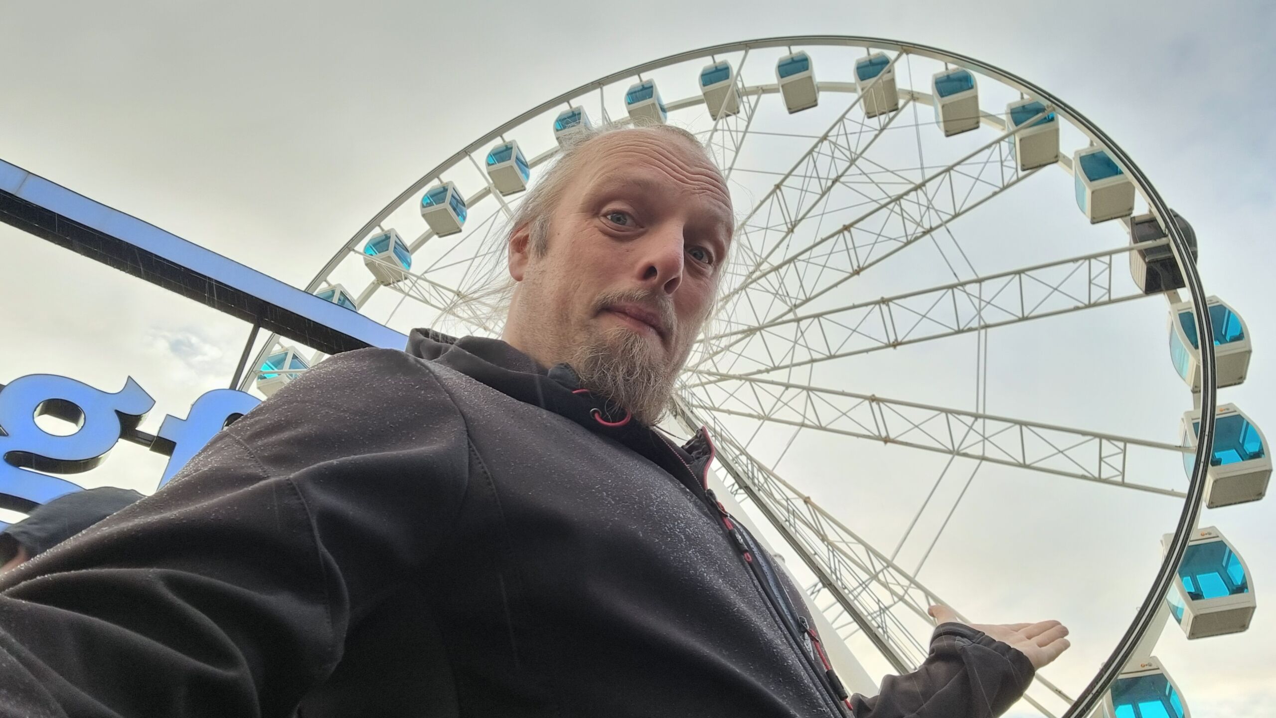 Full image of Dan stands in front of a ferris wheel against a cloudy sky.