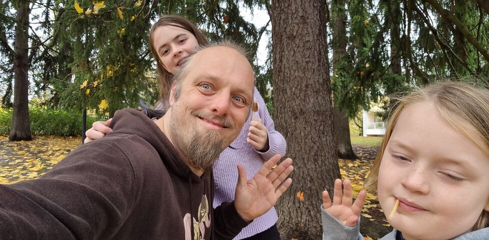 Dan and two children wave cheerfully from in front of a tree.