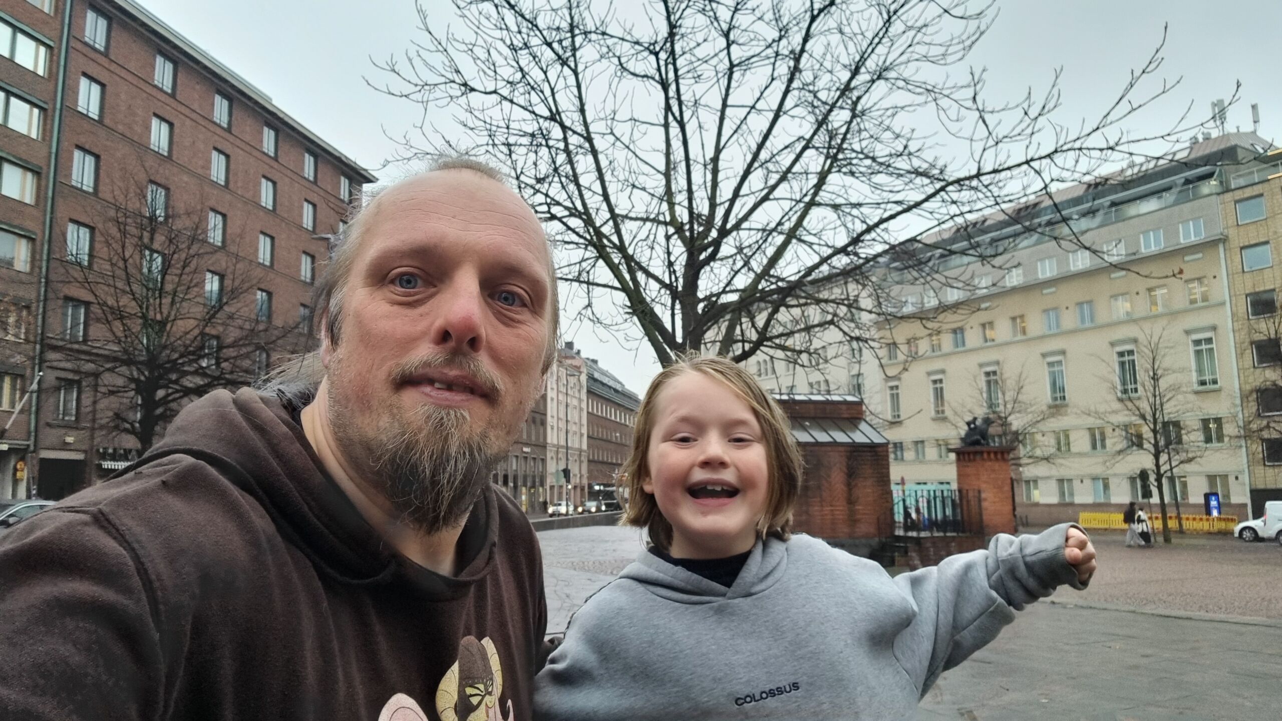 Full image of Dan and a young boy pose in front of a statue of two bears, atop a brick plinth in a city square.