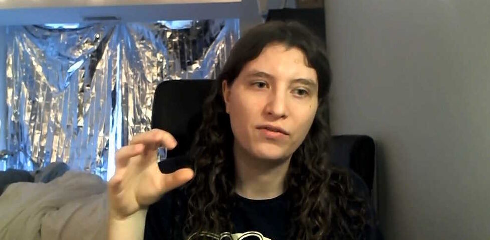 A white person with long black hair sits in a a room with fairy lights and a foil curtain, making a 'grabbing' gesture to the camera.