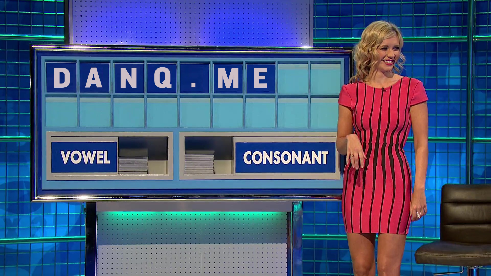 Full image of Rachel Riley, wearing a pink and black dress, stands in front of a partially-filled Countdown board showing the letters 'DANQ.ME'.