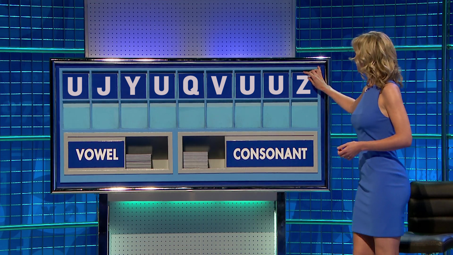 Full image of Rachel Riley, wearing a blue dress, places the last tile on a board to complete the tileset 'U J Y U Q V U U Z'.