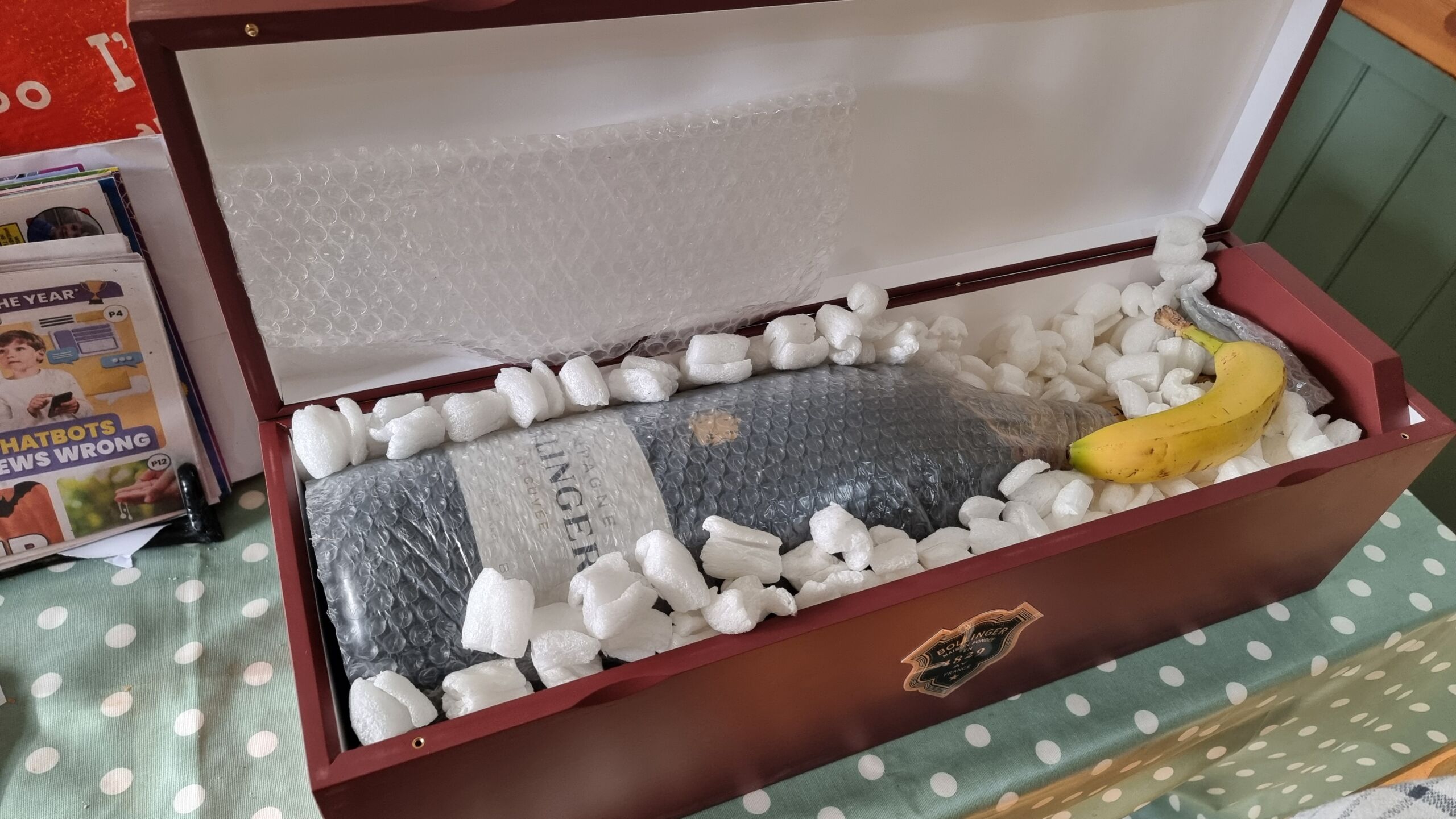 Full image of A six-litre bottle of champagne, wrapped in bubble wrap and surrounded by packing peanuts, in a wooden transport case, with a banana resting atop it.