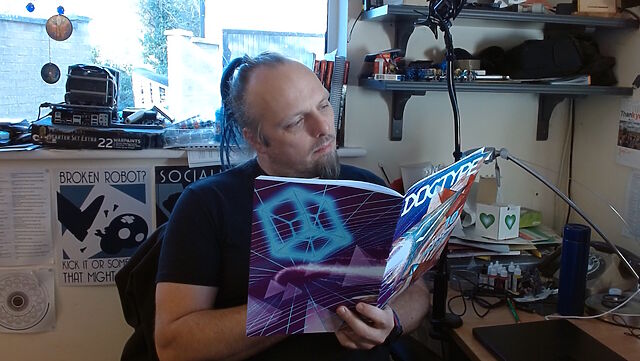 Dan sits at a cluttered desk reading a copy of DOCTYPE, a magazine with an aggressively 'cyberspace circa 1990' graphic design cover.