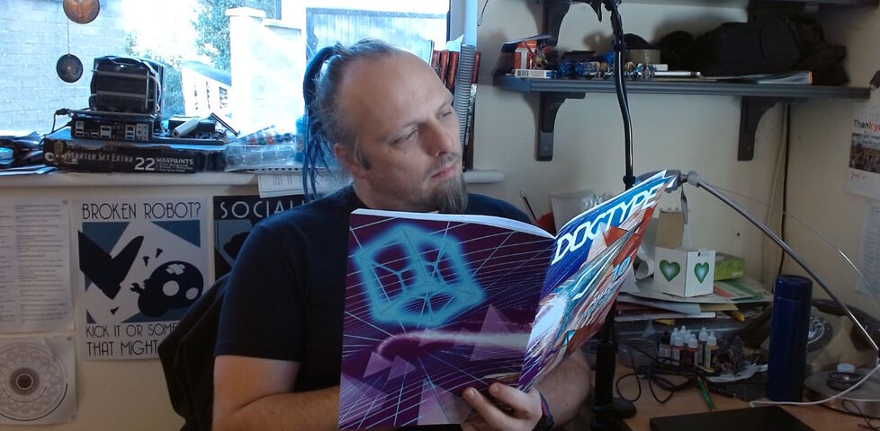 Dan sits at a cluttered desk reading a copy of DOCTYPE, a magazine with an aggressively 'cyberspace circa 1990' graphic design cover.