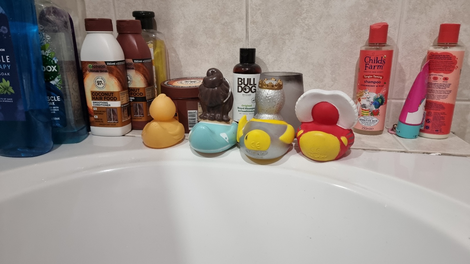 Full image of Four thematic rubber ducks sit along the edge of a fitted white bathtub, seemingly deliberate in their placement which sees them facing directly away from the bather and towards various shampoo bottles and a candle in a glass.