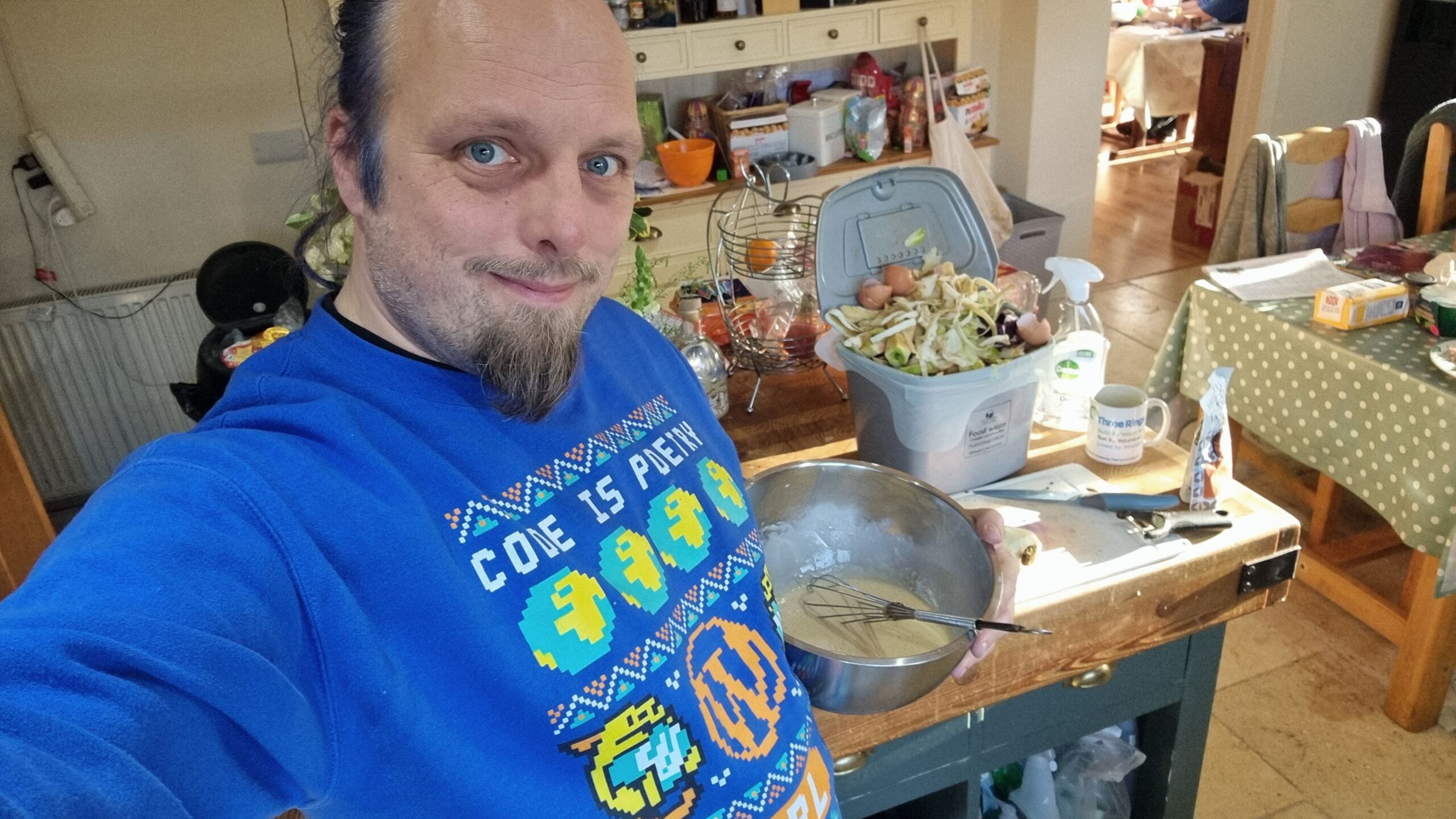Full image of Dan, a white man with a beard and blue hair, wearing a WordPress-themed Christmas jumper, beats a bowl of batter.