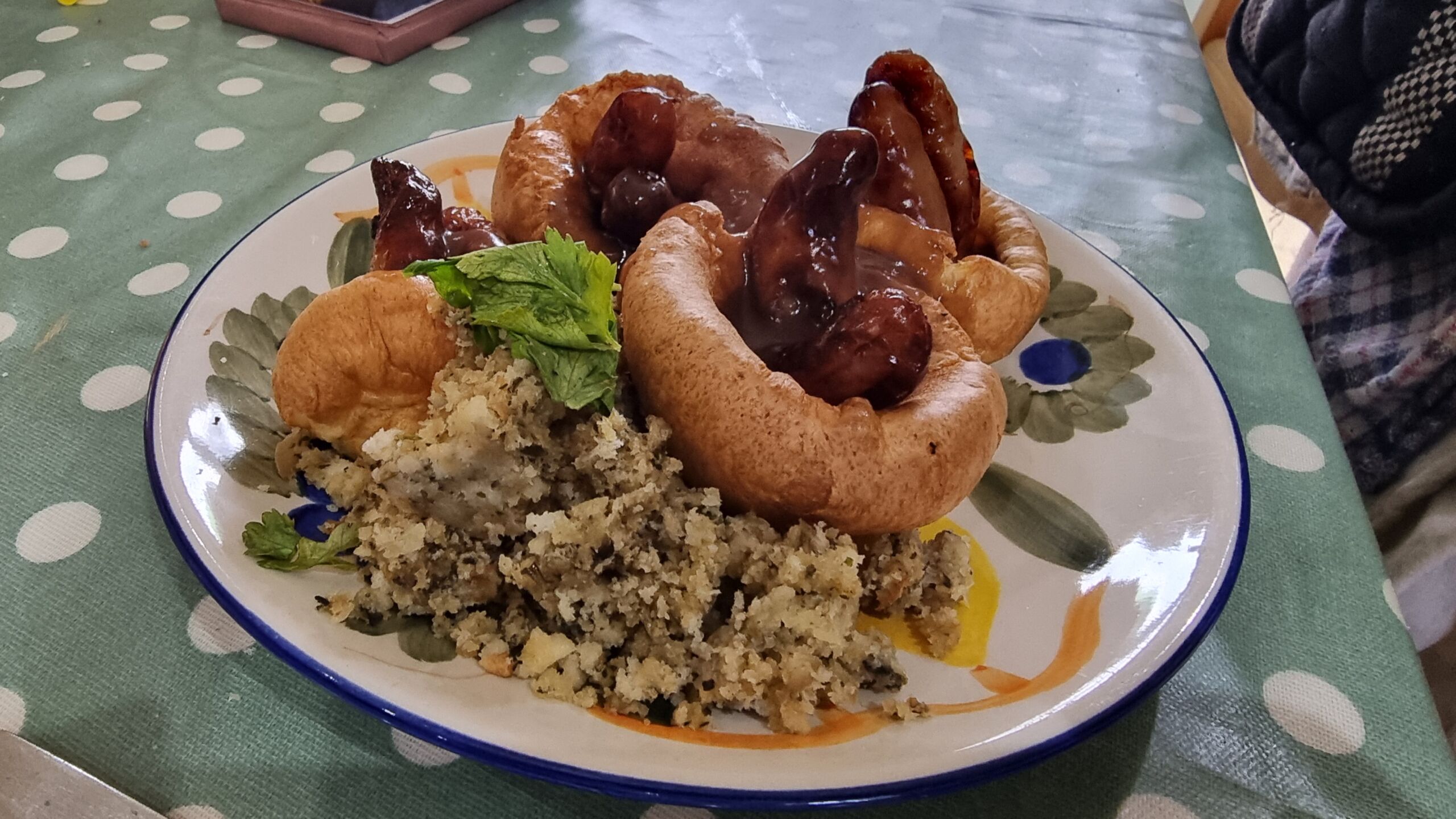 Full image of Four Yorkshire puddings on a side plate, each with one or two chipolata sausages sticking out, smothered in gravy, alongside a small quantity of sage & onion stuffing.