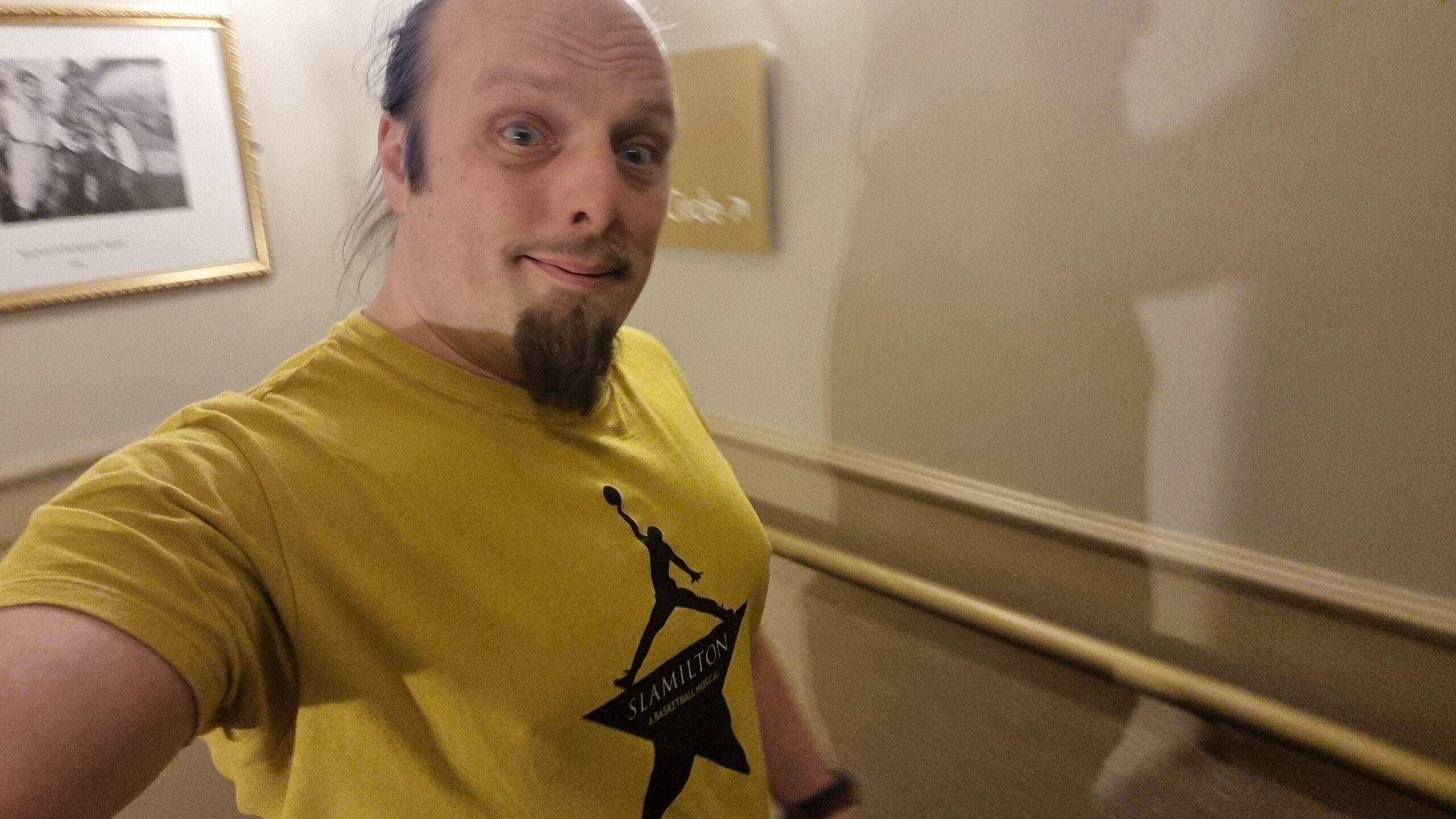 Full image of Dan, a white man with a goatee beard and a blue ponytail, wears a 'Slamilton' t-shirt in a theatre stairwell.